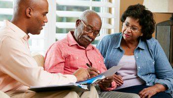 Financial Advisor Talking To Senior Couple At Home Signing Documents Sitting On Sofa